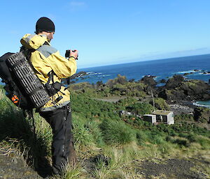 Expeditioner with back pack high on a slope looking down on a hut and the coast line
