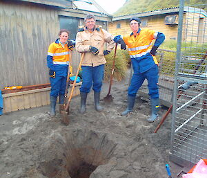 Three expeditioners standing next to a large hole holding shovels and picks