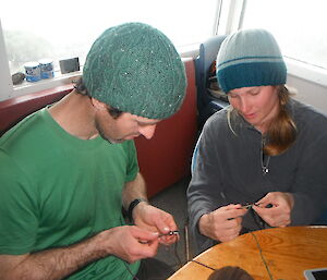 Female expeditioner instructs male hunter how to knit in a hut