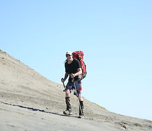 Expeditioner walking with a back pack and walking poles along a sandy hill