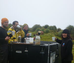 Four expeditioners standing around a large black pod drinking tea
