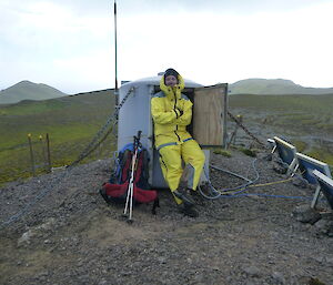 Expeditioner looking very cold leaning against a small hut, solar panels to his left