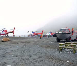 Three red and white helicopters with a LARC in the foreground on a gravel beach area