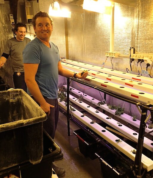 Aaron and Josh standing inside the hydroponics hut. There are two shelves with three rows of plastic hydroponic beds