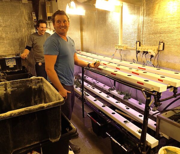 Aaron and Josh standing inside the hydroponics hut. There are two shelves with three rows of plastic hydroponic beds