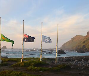 Three flags at half mast in commemoration of the first anniversary of the passing of Neil Adams. The first flag is green and yellow ANARE, the second is the Australian flag and the third is blue and white UN World Meteorological Organisation flag with Hasselborough Bay and North Head in the background