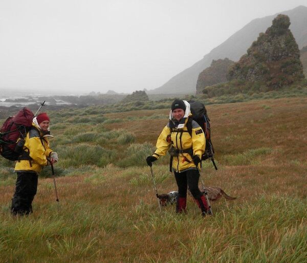 Ange and Leona standing in grass field, both with survival gear and backpacks and the rodent dogs near Leona’s feet. The hills in the background slope steeply toward the coast
