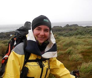 Leona standing in the tussock grass, wearing a yellow rain jacket and carrying a survival backpack. The coast is in the background