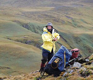 Tom standing on a slope with rolling hills and valleys in the background. Tom is wearing his yellow rain jacket, blue over pants and a pith helmet. He is standing beside two survival backpacks