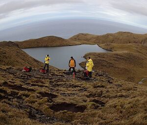 Clive, Marty and John standing in the field their packs are on the ground, behind them is a lake and then the ocean