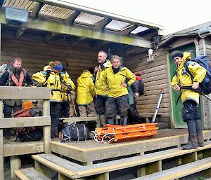 Team members outside on deck preparing for departure all are wearing their yellow jackets one team member has their back pack on and there is an orange stretcher placed on the deck