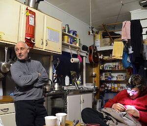 Inside Bauer Bay hut — Aaron (sitting) fixing a tear in his pants with dental floss and a piece of a guitar string while Tony leans with his back against the kitchen bench