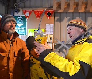 Marty, on the left holding up a piece of equipment (EPIRB) while Clive and Patty look on