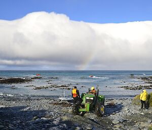 Looking out from Landing Beach, with the green tractor at the waters edge and two boats in the water, about 100 metres offshore. A cloud bank on the horizon with a part rainbow in the centre background