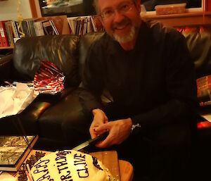 Dr Clive Stauss sitting down on a couch leaning forward ready to cut his birthday cake with the knife in hi hand