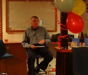 Tony finishing of his dinner sitting on a stool a white board behind him the bar on the right of him with some balloons and streamers attached to it