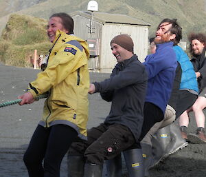 Leona, Steve, Dean, Nick, Mike and Billy pulling on a rope in the dark sand with a building and tussock grass in the back ground.