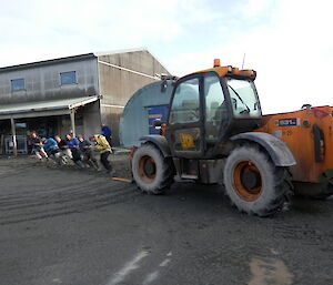 Kiwi team members pulling on a rope tied to a tractor on dark sand with the carpenters building in the back ground and tom leaning up against it.