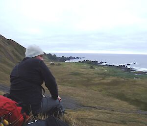 Josh resting during the hike for Mt Eitel to Bauer Bay, between jobs . He is sitting on a steep slope looking out to the view of the ocean. Is he enjoying the view or thinking for the moment about his normal job where he drives his ute between jobs