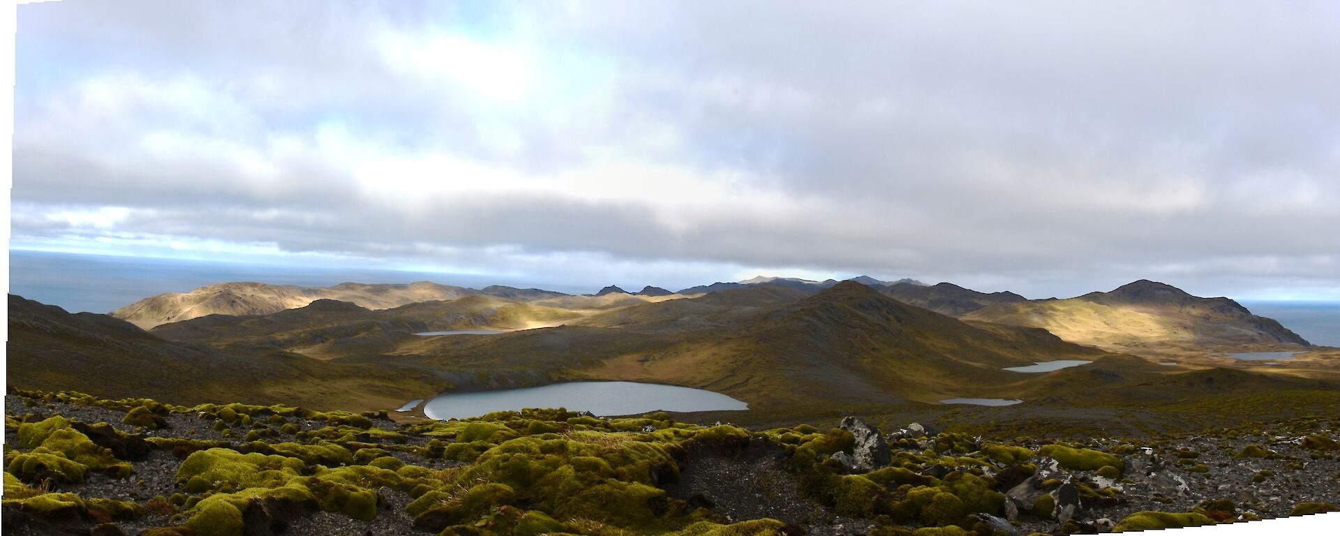 A panorama of the rugged mountainous landscape of Macquarie Island. Eroded hill tops covered in thin layers of cushion plants, the terrain disappearing to the south with the Southern Ocean on the distant horizon. Lake Eitel is in the foreground, one of the of many small fresh water lakes atop the main plateau