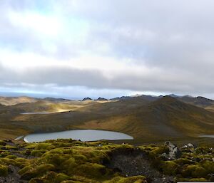 A panorama of the rugged mountainous landscape of Macquarie Island. Eroded hill tops covered in thin layers of cushion plants, the terrain disappearing to the south with the Southern Ocean on the distant horizon. Lake Eitel is in the foreground, one of the of many small fresh water lakes atop the main plateau