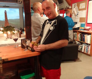 Tony standing at the bar with a blue mow hawk red boxers and a black tishirt holding a pair of darts in the background the Eifel Tower