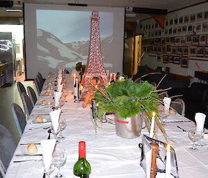 Table set with a white table clothe cuttlery, wine glasses, candles, the Eifel tower with bread leaning up against it and the a projection of the french alps on a screen in the background