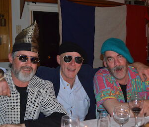 Clive wearing sunglasses and a silk hat , John wearing sunglasses and a beret and Greg wearing a very colourful shirt and beret all sitting at the table with the french flag behind them