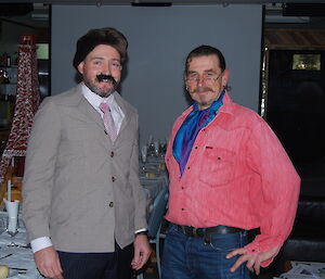 Craig dressed up in a jacket, fake mustache and a fury hat and Billy dressed in a pink shirt with a blue cravat in the background is a screen and to the left of graig is the replica Eifel tower in the background