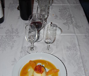 A photo of a desert called creme caramel with sweet sauce and orange pieces set on the table behind the plate are some wine glasses