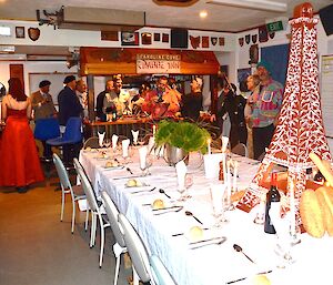 The table is with white table clothe, cuttlery, wine glasses, candles and in the middle is a small version of the Eifel tower with french bread leaning up against in in the background everybody is gathered at the bar dressed in their best attire