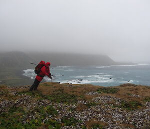 Dave wearing his red jacket and backpack at standing leaning on his walking pole at the top of North head with a view of Macquarie island with very low laying clouds and the ocean in the background.