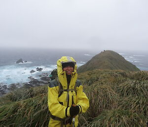 Patty in her yellow jacket holding her walking pole standing amongst tussock grass. In the back ground is the northern peninsula of the island the ocean and cloud cover