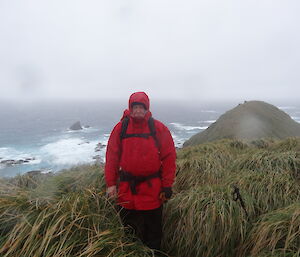 Dave stands frowning amongst the tussock grass holding his walking pole in the back is the north Peninsula of Macquarie Island the ocean and low laying cloud