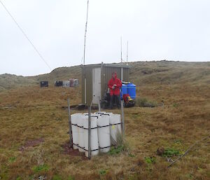 David in his red jacket is holding his gloves he is standing next to the radio shack his bag is resting on a blue water tank the sack has antennas and in the back ground there are some cargo bins. In the forefront is a square made up of piping used to grow a specific plant for science.