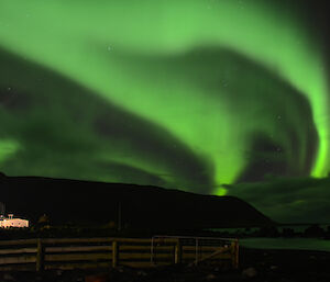 Green tinged aurora in the western sky, with the island hills silhouetted and the science building lit up in the bottom left. There is a greenish reflection ioff the low clouds and the waters of Hasselborough Bay