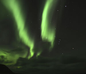 Two green tinged aurora ‘curtains’ in the western sky, with the island hills silhouetted in the bottom left. There is a greenish reflection off the low clouds. Many stars can be seen in the sky including the ‘big dipper'