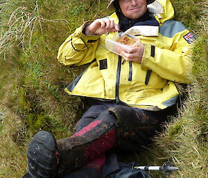 Tom wearing his floppy hat and yellow jacket, sitting down with his feet proped up on his bags while he eats lunch