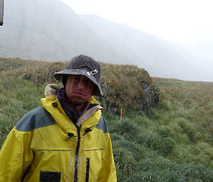 Tom wearing a hat looking unhappy and wet standing outside in the rain. In the background is tussock and a mountainin