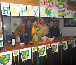 Mark with his arm around Chef Tony, Dave and Greg behind the bar that is decorated in the Norwich city football logo and green and yellow letters that say happy birthday
