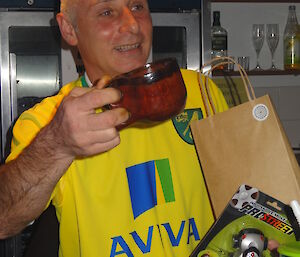 Chef Tony in his yellow Norwich city football jersey holding the handcrafted wooden mug Dave made for him and in the other hand a gift bag and his toy car.