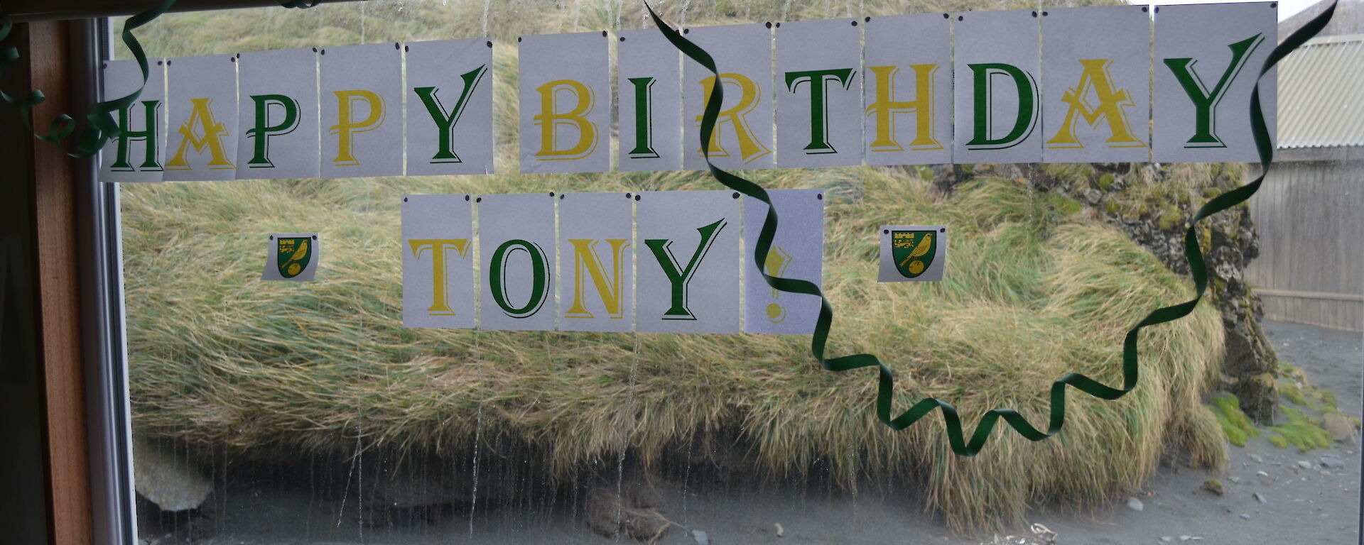 Happy Birthday Tony printed out in green and yellow and stuck onto the Mess window outside there is a mound of tussock and dark sand