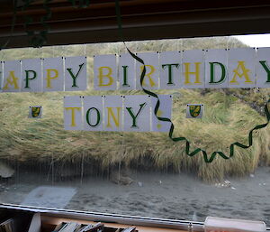 Happy Birthday Tony printed out in green and yellow and stuck onto the Mess window outside there is a mound of tussock and dark sand
