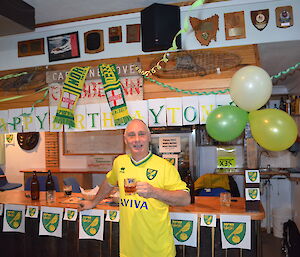 Chef Tony standing in front of the bar that is decorated in the Norwich city football logo and green and yellow letters that say happy birthday