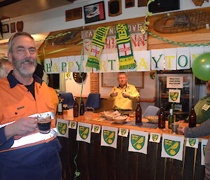 Greg in the forefront withtony behind him, Dave in the background behind the bar, Dr Clive sitting behind the bar and Chris next to the bar that is decorated in the Norwich city football logo and green and yellow letters that say happy birthday Tony