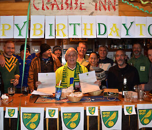All the crew behind the bar that is decorated in the Norwich city football logo and green and yellow letters that say happy birthday