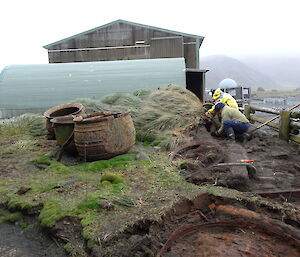 Patty and Chris looking down into the ground to see what they’re uncovering in the forefront there is an old boiling pot full of water in the background you can see the ocean