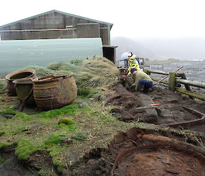 Patty and Chris excavating uncovering artefacts the area is made up of uneven ground tussock and there are 3 large boiling pots sitting to one side full of water in the background you can see the ocean