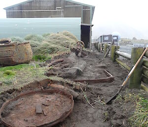 Excavation uncovering artefacts the area is made up of uneven ground tussock and there are 3 large boiling pots sitting to one side full of water in the background you can see the ocean