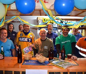 Everybody celebrating behind the bar that’s decorated in balloons and streamers coloured yellow and blue craig is sitting in the middle